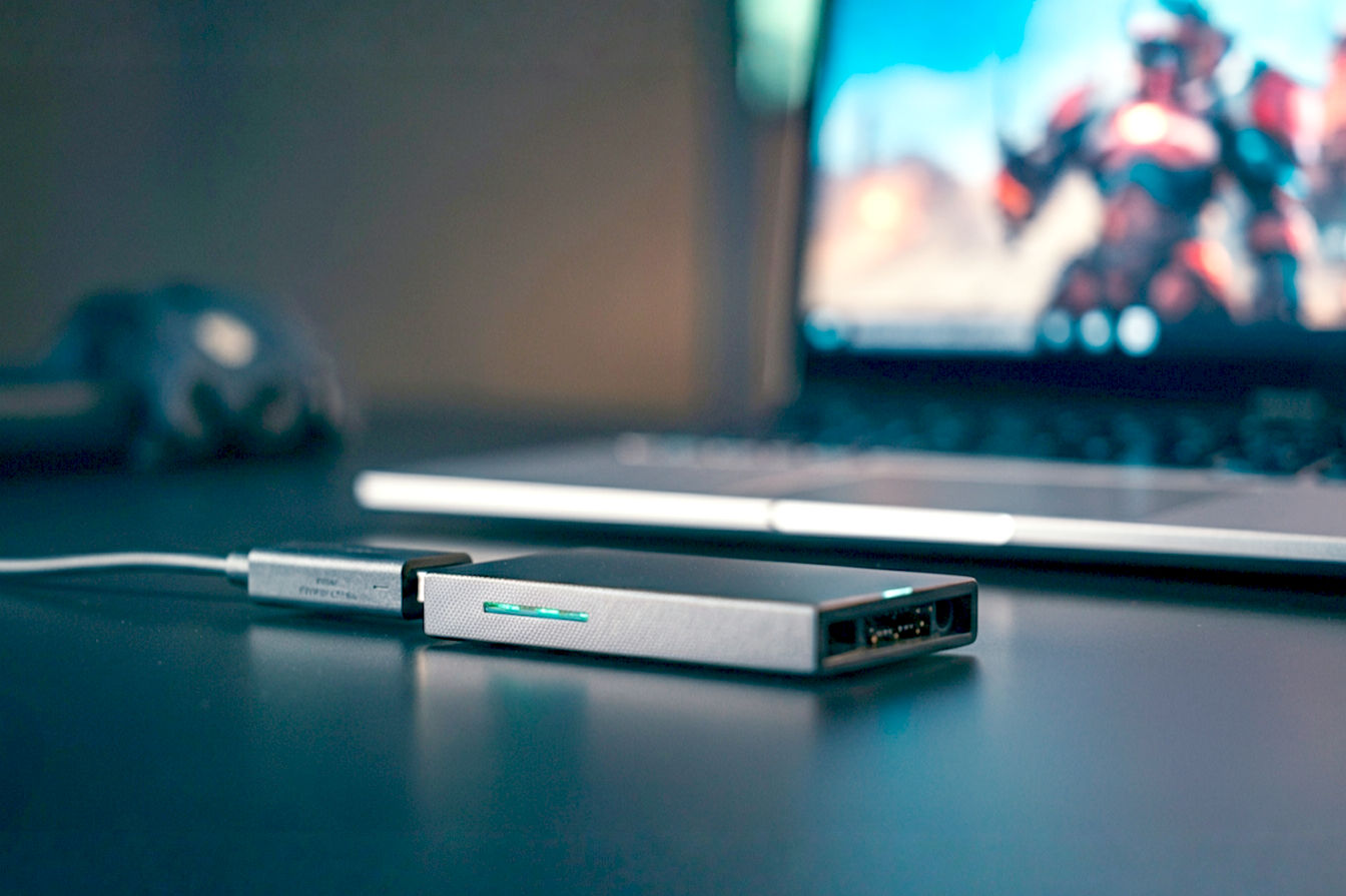 Professional, high-angle photograph showing a sleek, modern external USB SSD connected to a slim gaming laptop. The laptop screen displays a vibrant game on screen. The background is a slightly blurred, modern desk or travel setting, emphasizing portability and technology. Good lighting highlights the textures of the devices.