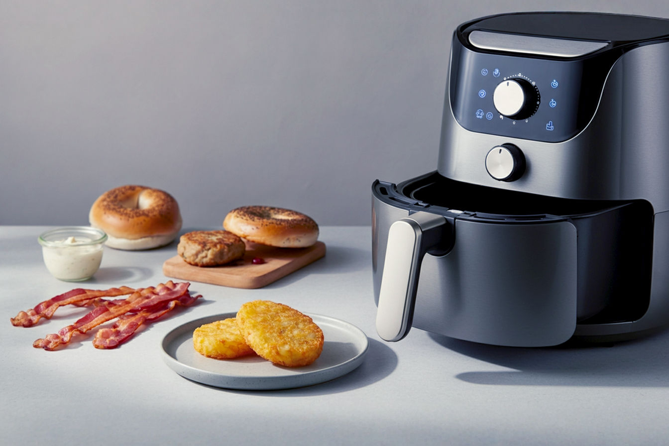 A beautifully styled, high-resolution food photograph on a clean, modern kitchen counter. In the foreground, a compact, sleek air fryer (e.g., matte black or stainless steel) is subtly visible. Artfully arranged around it are small, appealing portions of various air-fried breakfast items