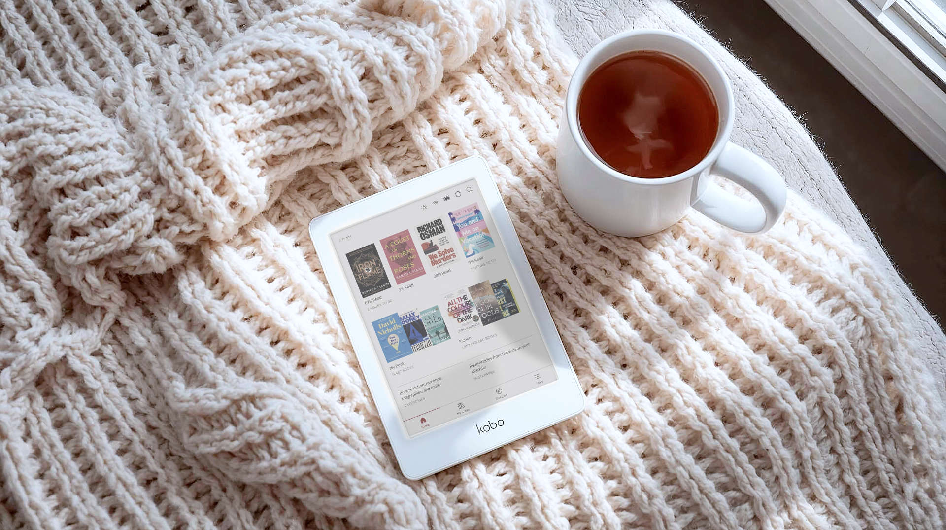 A high-resolution, top-down shot of the Kobo Clara Colour e-reader lying on a cozy knitted cream blanket next to a steaming mug of tea. The screen displays a vibrant, colourful comic book cover. Soft natural lighting from a nearby window illuminates the texture of the blanket and the matte finish of the screen.