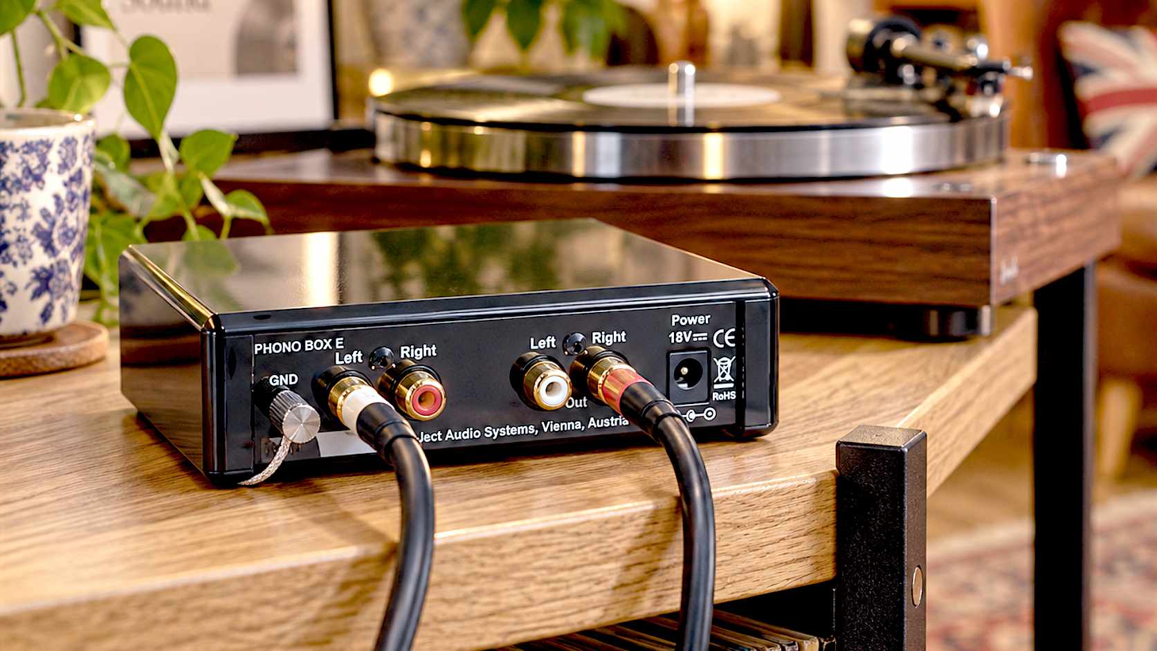 A hyper-realistic, beautifully lit photograph of a glossy black Pro-Ject Phono Box E sitting on a solid oak hi-fi rack. In the softly blurred background, a high-end vinyl record player is spinning a record. Warm, ambient lighting highlights the gold-plated RCA cables connecting the units, evoking a cozy British listening room.