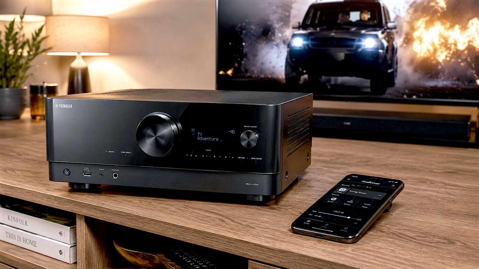 A hyper-realistic, warmly lit photograph of the sleek, black Yamaha RX-V6A receiver sitting on an oak media unit in a cosy, modern British living room. A glowing TV screen displaying an action movie is blurred softly in the background, and a smartphone showing the MusicCast app rests on the unit nearby.